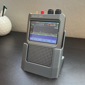 Puede incluir: Un receptor de radio Malahit DSP2 gris con una pantalla digital que muestra una frecuencia de 14.160.000 MHz. La radio está sentada en un soporte gris impreso en 3D.