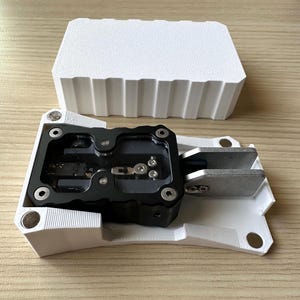 May include: A white and black mechanical device with a rectangular white block in the background. The device has a complex design with multiple screws and metal components. The block has a textured surface.