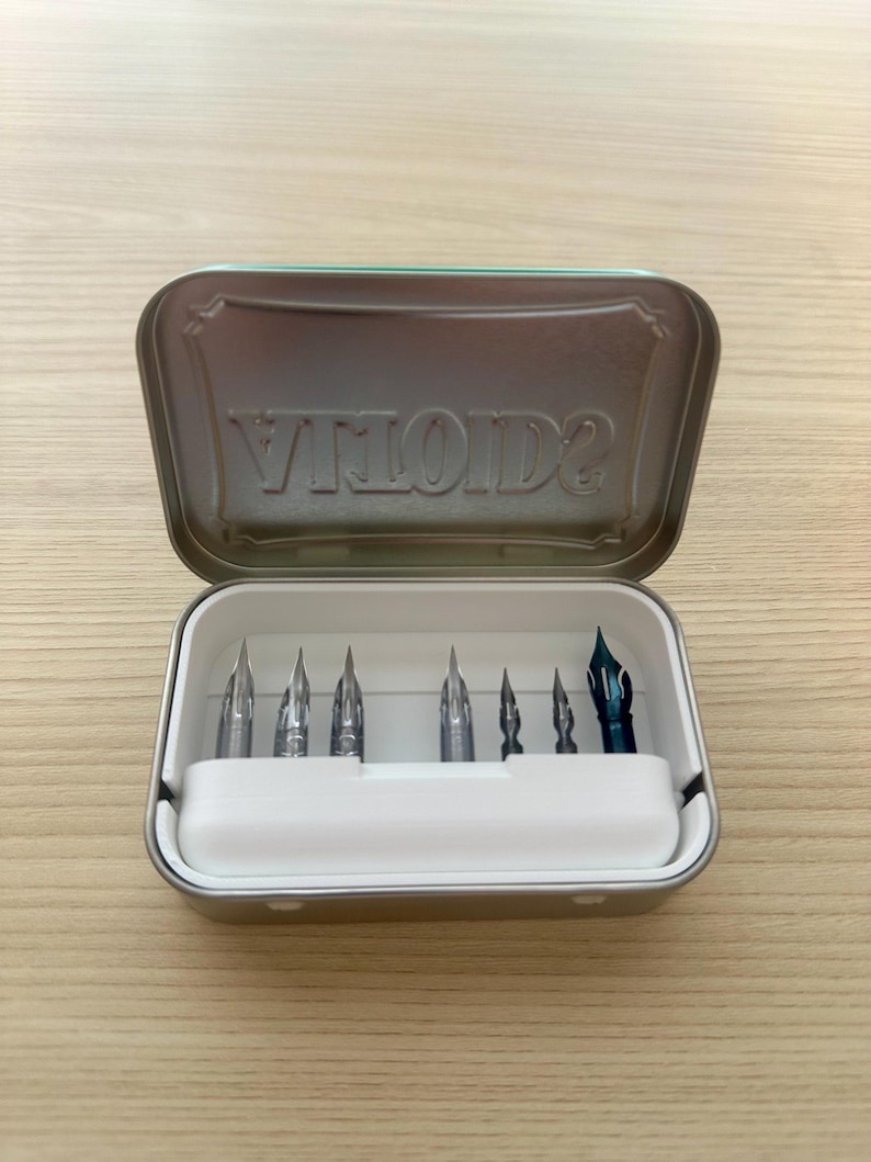 May include: A set of calligraphy pen nibs in a metal tin. The tin is open, revealing several silver nibs and one blue nib. The tin is light green with the word "VILOIDS" embossed on the inside of the lid.