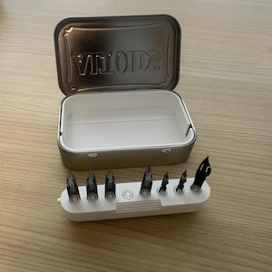 May include: An open, rectangular metal tin with the word "VINTAGE" embossed on the lid. A white plastic holder contains several black pen nibs. The tin and nib holder are on a wooden surface.