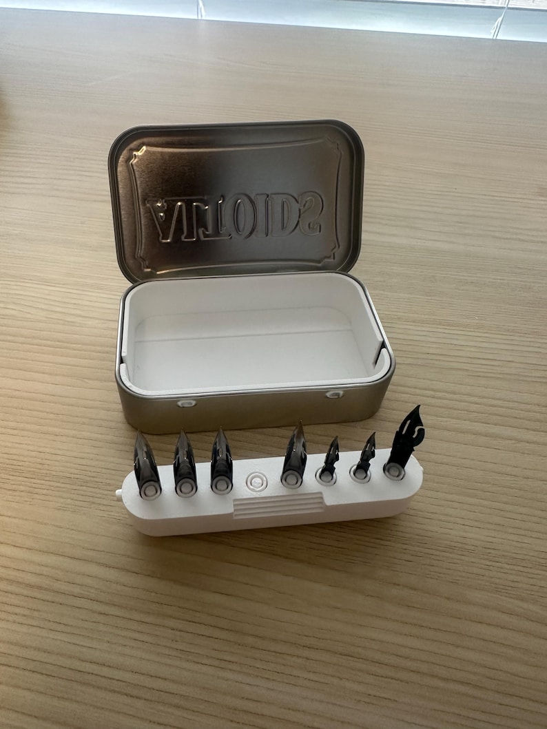 May include: An open, rectangular metal tin with the word "VINTAGE" embossed on the lid. A white plastic holder contains several black pen nibs. The tin and nib holder are on a wooden surface.