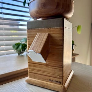 May include: A wooden NutriMill grain mill with a bamboo exterior and a brown bowl on top. The mill has a white chute and is sitting on a light-colored surface. The text "NutriMill" is printed on the front.