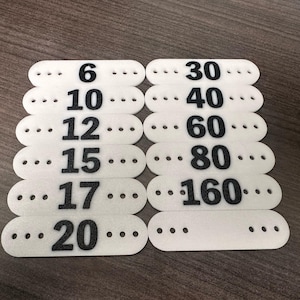 Può includere: Tessere numeriche in schiuma bianca con numeri neri: 6, 10, 12, 15, 17, 20, 30, 40, 60, 80 e 160. Ogni tessera ha tre piccoli fori. Le tessere sono disposte in due colonne.