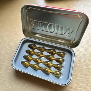 May include: A rectangular metal tin with a hinged lid embossed with the word ALTOIDS. Inside, a custom grey 3D-printed insert holds ten brass-coloured bullet-shaped objects arranged in five pairs, each featuring a rounded tip and a flat base.