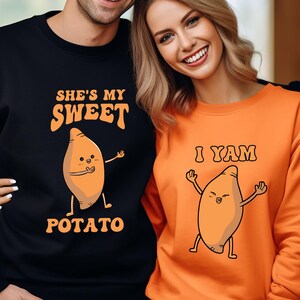 Puede incluir: Sudaderas negras y naranjas a juego. La sudadera negra dice "She's my Sweet Potato" con una batata de dibujos animados. La sudadera naranja dice "I Yam" con una batata de dibujos animados. La pareja sonr&iacute;e.