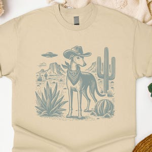 May include: A beige t-shirt featuring a desert scene with a dog wearing a cowboy hat and bandana. The dog stands in front of cacti and desert plants. The graphic is in shades of blue and gray.