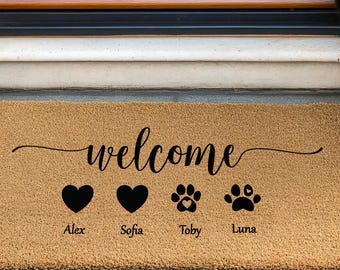 May include: A rectangular coir doormat with the word "welcome" in a cursive font. Below the word are two black hearts and two paw prints, each with a name underneath. The mat is tan and sits on a gray surface.