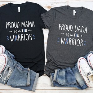 May include: Two t-shirts, one black and one gray, with the text "Proud Mama of a TID Warrior" and "Proud Dada of a TID Warrior" on them. Both shirts feature a blue and gray ribbon.