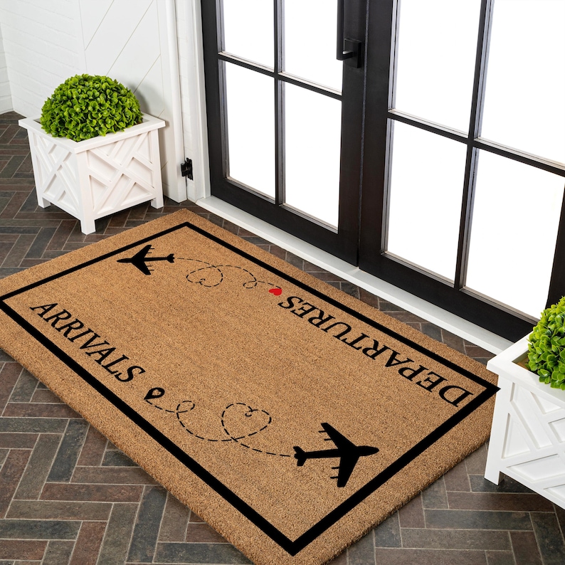 May include: A rectangular doormat with a brown coir surface and a black border. The mat features the words "ARRIVALS" and "DEPARTURES" in black, along with airplane silhouettes and dotted flight paths with hearts. A decorative welcome mat for a home.