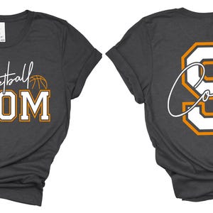 May include: Dark gray t-shirt with "Basketball MOM" in white and gold lettering, a basketball graphic, and the name "Coleen" and the number 9 on the back. The shirt has a crew neck and short sleeves.