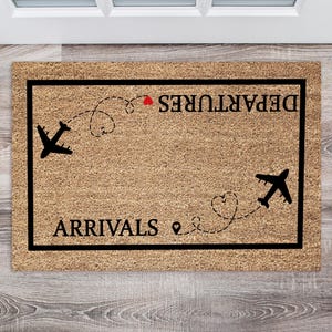 May include: A rectangular doormat with a brown coir surface and a black border. The mat features the words "ARRIVALS" and "DEPARTURES" in black, with airplane silhouettes and dotted flight paths. A red heart is above the word "DEPARTURES".