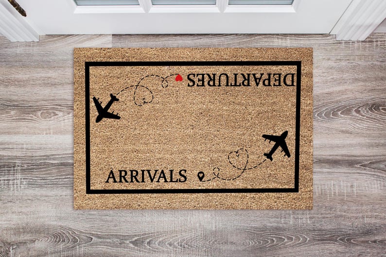 May include: A rectangular doormat with a brown coir surface and a black border. The mat features the words "ARRIVALS" and "DEPARTURES" in black, with airplane silhouettes and dotted flight paths. A red heart is above the word "DEPARTURES".