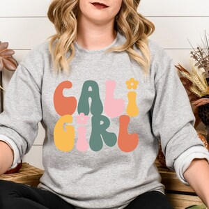 K&ouml;nnte beinhalten: Ein graues Sweatshirt mit dem Text "CALI GIRL" in bunten, Retro-Lettern. Die Buchstaben sind schwarz umrandet und haben einen leicht erhabenen 3D-Effekt.
