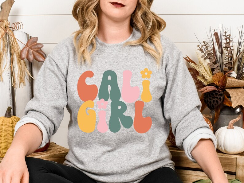 K&ouml;nnte beinhalten: Ein graues Sweatshirt mit dem Text "CALI GIRL" in bunten, Retro-Lettern. Die Buchstaben sind schwarz umrandet und haben einen leicht erhabenen 3D-Effekt.
