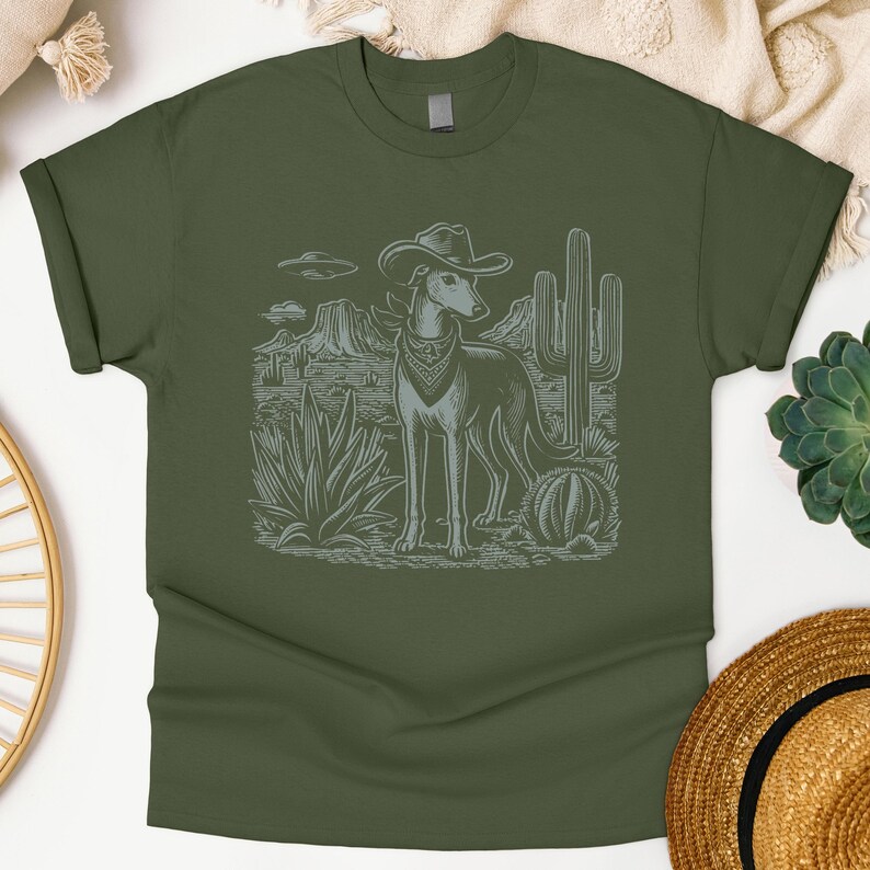May include: Olive green t-shirt featuring a desert scene with a greyhound wearing a cowboy hat and bandana. The graphic includes cacti, mountains, and a UFO. The shirt is made of soft material.