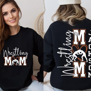 以下が含まれることがあります： 黒色のスウェットシャツ。白の筆記体とブロック体で「Wrestling Mom」の文字とレスリングのグラフィックがプリントされています。スウェットシャツの背面には「Wrestling Mom」の文字と「Jackson」の名前がプリントされています。