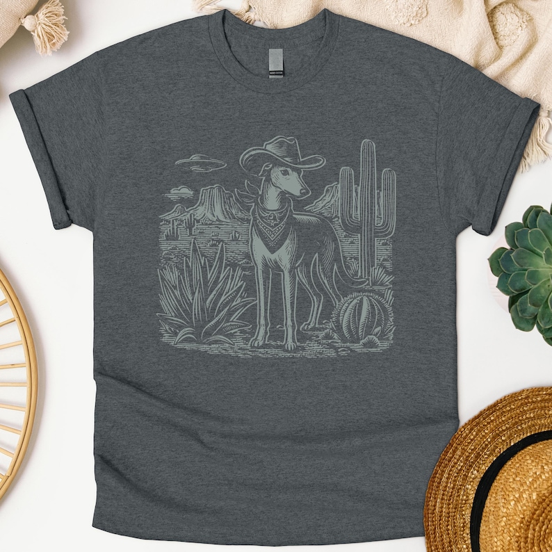 May include: Dark gray t-shirt featuring a detailed illustration of a dog wearing a cowboy hat and bandana, set against a desert landscape with cacti and mountains. The design is in a light gray color.