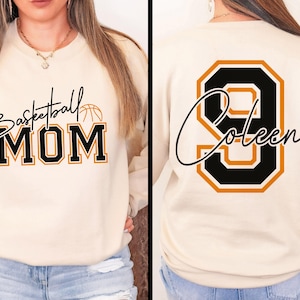 May include: Cream-colored sweatshirt with the text "Basketball MOM" in black and gold lettering, with a basketball graphic. The back of the sweatshirt features the number "9" and the name "Coleen" in a similar style.