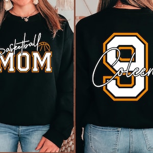 May include: Black sweatshirt with "Basketball MOM" in white and gold lettering on the front. The back features a large white number 9 with gold trim and the name "Coleen" in white script. The sweatshirt is worn with blue jeans.
