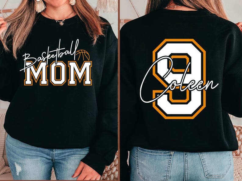 May include: Black sweatshirt with "Basketball MOM" in white and gold lettering on the front. The back features a large white number 9 with gold trim and the name "Coleen" in white script. The sweatshirt is worn with blue jeans.