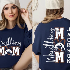 以下が含まれることがあります： ネイビーブルーのスウェットシャツ。白の筆記体とブロック体で「Wrestling MOM」の文字とレスリングのグラフィックが描かれています。シャツの背面には、白で「Wrestling MOM」と「Jackson」の文字が書かれています。