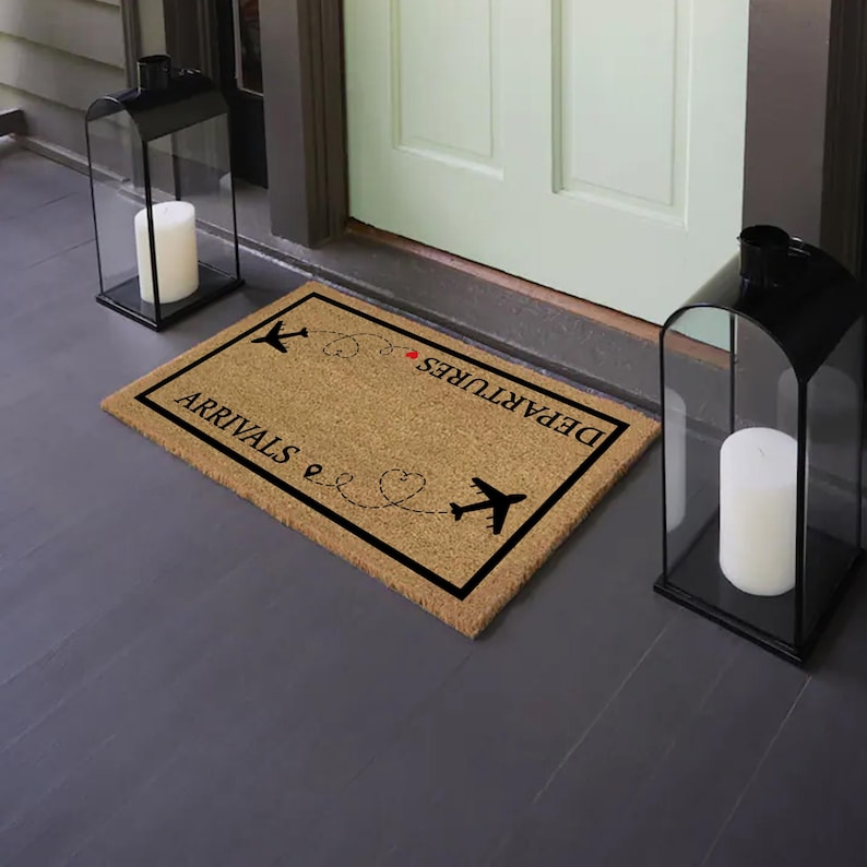 May include: A rectangular doormat with the words "ARRIVALS" and "DEPARTURES" in black, along with airplane illustrations and a heart-shaped design. The mat is made of a natural fiber material and has a black border. Two black lanterns with white candles flank the mat.