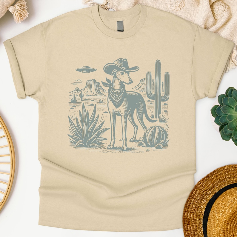 May include: A beige t-shirt featuring a desert scene with a dog wearing a cowboy hat and bandana. The dog stands in front of cacti and desert plants. The graphic is in shades of blue and gray.