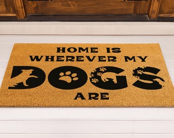 May include: Rectangular doormat made of natural coir fiber. The mat features the phrase "HOME IS WHEREVER MY DOGS ARE" in black block letters. The word "DOGS" is creatively designed with dog silhouettes and paw prints.