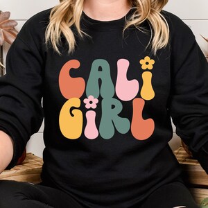 K&ouml;nnte beinhalten: Schwarzer Pullover mit einer Retro-Grafik, die "Cali Girl" in bunten, wellenf&ouml;rmigen Buchstaben mit kleinen Blumenakzenten sagt.