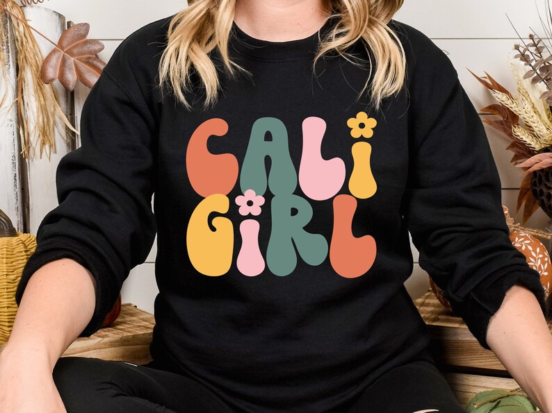 K&ouml;nnte beinhalten: Schwarzer Pullover mit einer Retro-Grafik, die "Cali Girl" in bunten, wellenf&ouml;rmigen Buchstaben mit kleinen Blumenakzenten sagt.