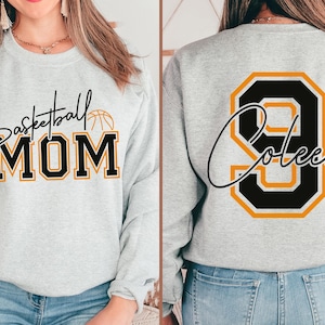 May include: Light gray sweatshirt with "Basketball MOM" in black and gold lettering. The back features a large black and gold number 9 with the name "Coleen" in script. A basketball graphic is above the word "MOM".