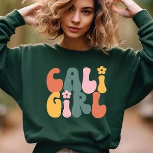 K&ouml;nnte beinhalten: Ein dunkelgr&uuml;ner Pullover mit dem Text "CALI GIRL" in bunten, Retro-Lettern. Die Buchstaben sind wei&szlig; umrandet und haben ein Blumenmuster.