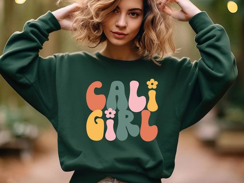 K&ouml;nnte beinhalten: Ein dunkelgr&uuml;ner Pullover mit dem Text "CALI GIRL" in bunten, Retro-Lettern. Die Buchstaben sind wei&szlig; umrandet und haben ein Blumenmuster.