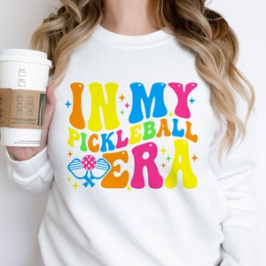 以下が含まれることがあります： カラフルなレトロなフォントで「In My Pickleball Era」と書かれた白いスウェットシャツ。テキストの下にピクルボールのパドルが描かれています。