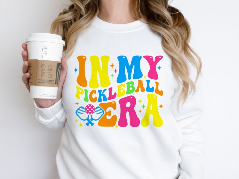 以下が含まれることがあります： カラフルなレトロなフォントで「In My Pickleball Era」と書かれた白いスウェットシャツ。テキストの下にピクルボールのパドルが描かれています。