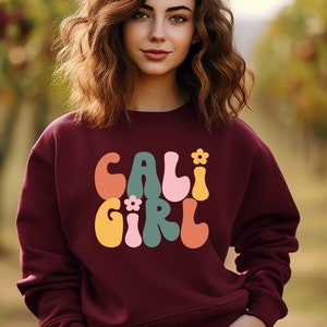 K&ouml;nnte beinhalten: Ein weinroter Pullover mit einer Retro-Grafik, die "CALI GIRL" in bunten, wellenf&ouml;rmigen Buchstaben mit kleinen Blumenakzenten zeigt.