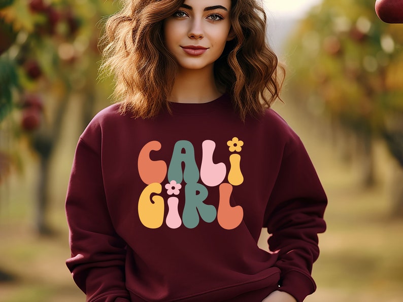 K&ouml;nnte beinhalten: Ein weinroter Pullover mit einer Retro-Grafik, die "CALI GIRL" in bunten, wellenf&ouml;rmigen Buchstaben mit kleinen Blumenakzenten zeigt.