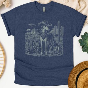 May include: Navy blue t-shirt featuring a desert scene with a dog wearing a cowboy hat and bandana. The graphic includes cacti, mountains, and an unidentified flying object. The design is in a muted, vintage style.