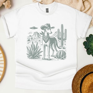 May include: White t-shirt featuring a desert scene with a greyhound wearing a cowboy hat and bandana. The design includes cacti, mountains, and a UFO. The graphic is in a monochromatic style.