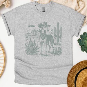 May include: A light gray t-shirt featuring a desert scene with a dog wearing a cowboy hat and bandana. The graphic includes cacti, mountains, and a UFO. The dog is the central focus of the design.