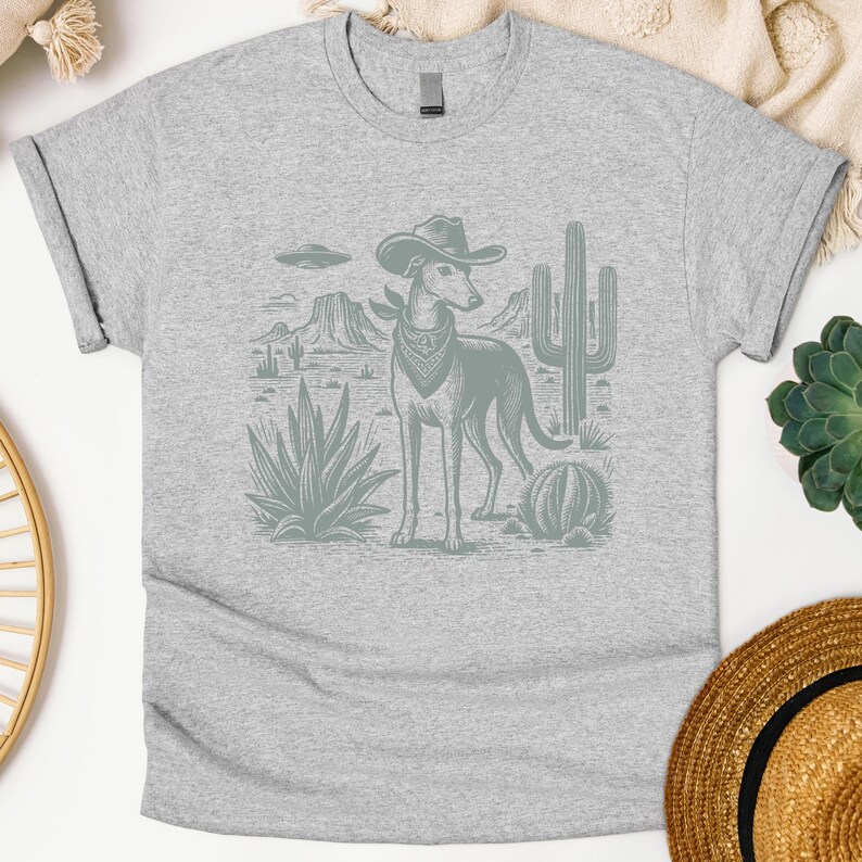 May include: A light gray t-shirt featuring a desert scene with a dog wearing a cowboy hat and bandana. The graphic includes cacti, mountains, and a UFO. The dog is the central focus of the design.