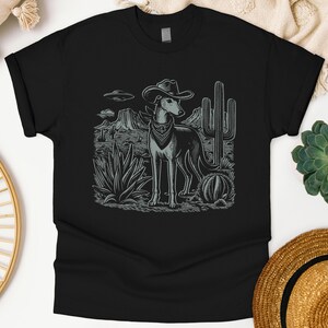May include: Black t-shirt featuring a grey illustration of a dog wearing a cowboy hat and bandana, set against a desert landscape with cacti and mountains. The design includes an image of a UFO in the sky.