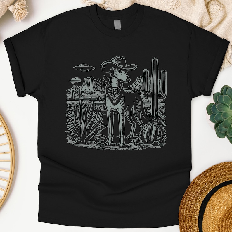 May include: Black t-shirt featuring a grey illustration of a dog wearing a cowboy hat and bandana, set against a desert landscape with cacti and mountains. The design includes an image of a UFO in the sky.