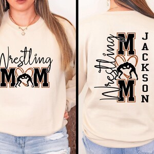 以下が含まれることがあります： クリーム色のスウェットシャツ。黒い筆記体とブロック体の文字で「Wrestling Mom」と書かれており、レスリングのフィギュアが入ったハート型のデザインが特徴です。スウェットシャツの背面には同じデザインと「Jackson」という名前があります。