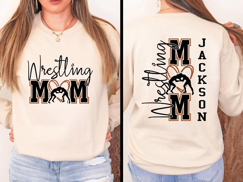 以下が含まれることがあります： クリーム色のスウェットシャツ。黒い筆記体とブロック体の文字で「Wrestling Mom」と書かれており、レスリングのフィギュアが入ったハート型のデザインが特徴です。スウェットシャツの背面には同じデザインと「Jackson」という名前があります。