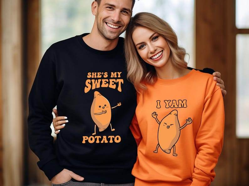 Puede incluir: Sudaderas negras y naranjas a juego. La sudadera negra dice "She's my Sweet Potato" con una batata de dibujos animados. La sudadera naranja dice "I Yam" con una batata de dibujos animados. La pareja sonr&iacute;e.