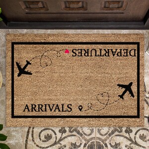 May include: A rectangular doormat with a black border and a light brown surface. The mat features the words "DEPARTURES" and "ARRIVALS" in black, along with airplane silhouettes and dotted flight paths. A small red heart is also included.