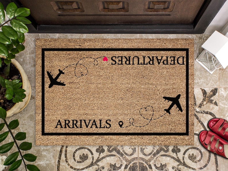 May include: A rectangular doormat with a black border and a light brown surface. The mat features the words "DEPARTURES" and "ARRIVALS" in black, along with airplane silhouettes and dotted flight paths. A small red heart is also included.