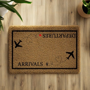May include: A rectangular brown doormat with black text that reads "ARRIVALS" and "DEPARTURES" with airplane graphics. The mat is surrounded by a black border. Two white sneakers are next to the mat on a wooden floor.