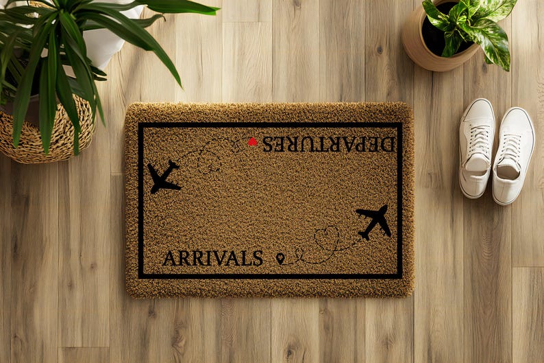May include: A rectangular brown doormat with black text that reads "ARRIVALS" and "DEPARTURES" with airplane graphics. The mat is surrounded by a black border. Two white sneakers are next to the mat on a wooden floor.